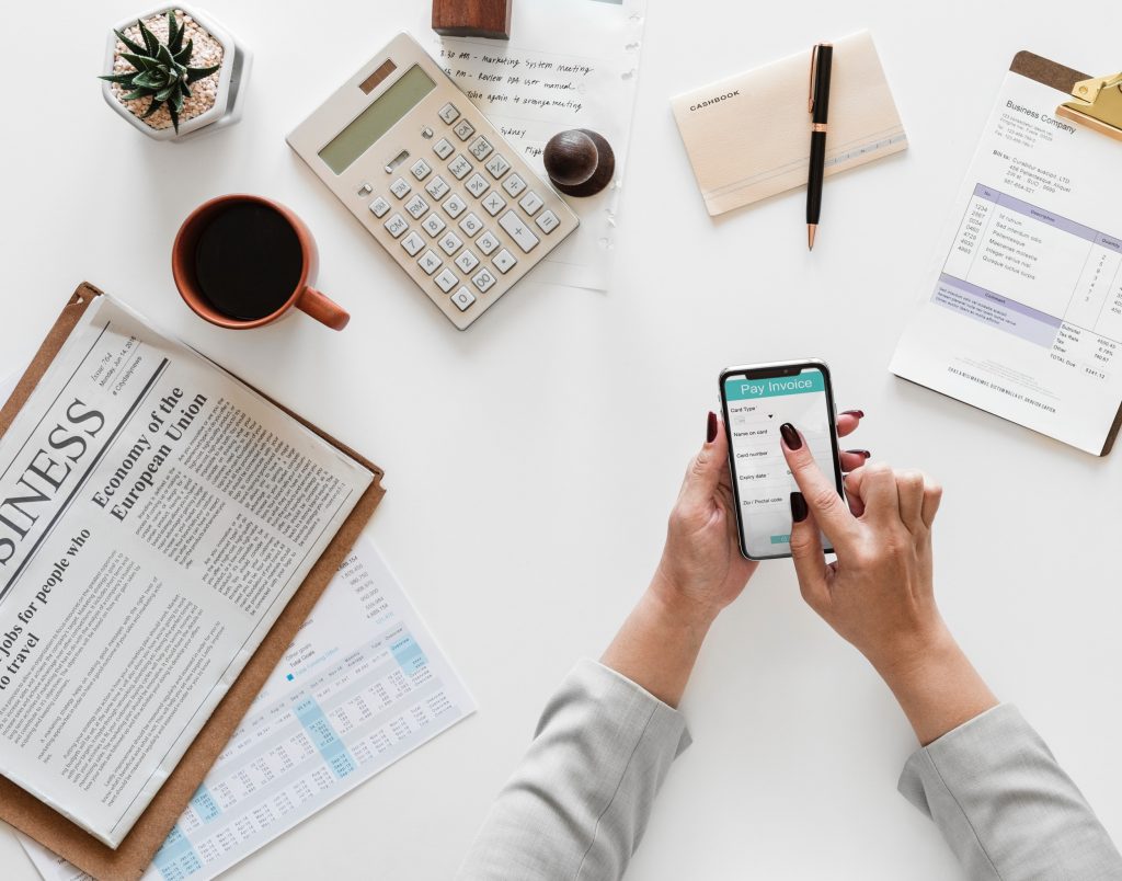 Uma mão manuseando um smartphone sobre uma mesa. na mesa estão uma xícara de café, um jornal, uma calculadorae algumas planinhas de planejamento. A imagem representa os pontos ee relevar ao criar uma Sociedade Simples de Advogados