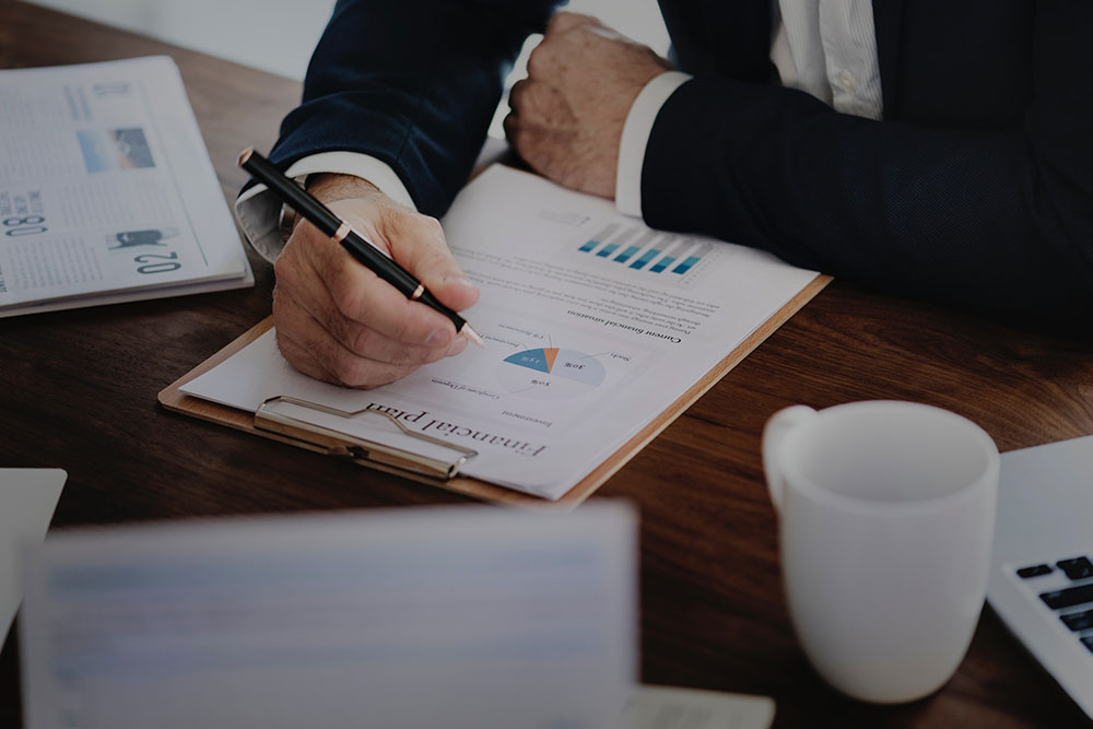 Homem deterno debruçado sobre uma mesa fazendo anotações com uma caneta em um papel contendo gráficos. Sobre a mesa, um teclado de computador e uma xícara de café. Imagem representa os cuidados financeiros a serem tomados em prol do sucesso do seu escritório
