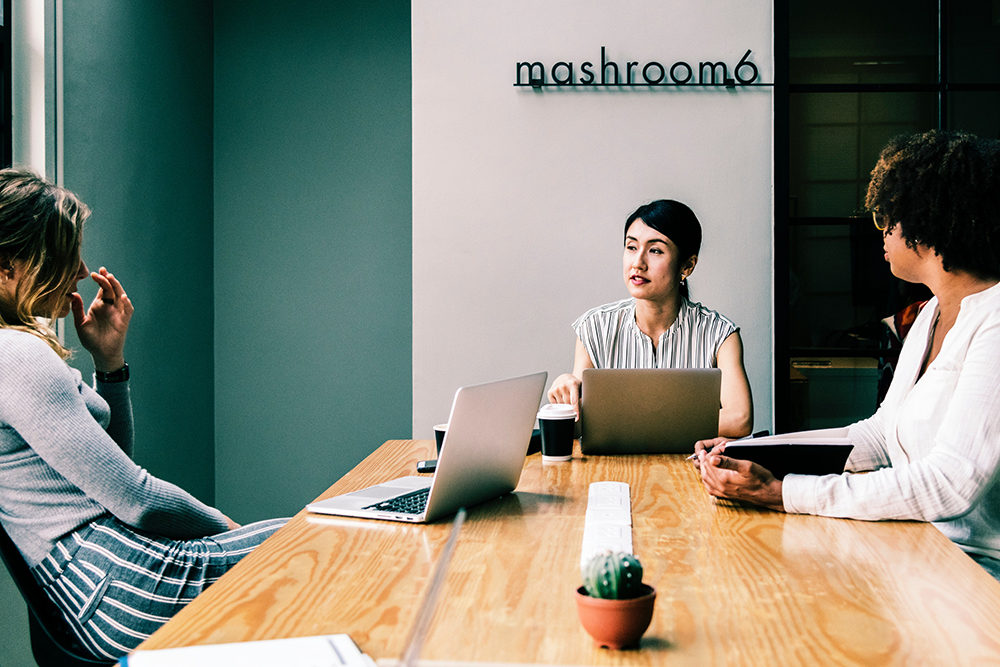 Três mulheres em reunião em uma mesa. Sobre a mesa há notebooks, copos de café e um cacto. Foto representa a melhoria em produtividade quando a equipe está conectada