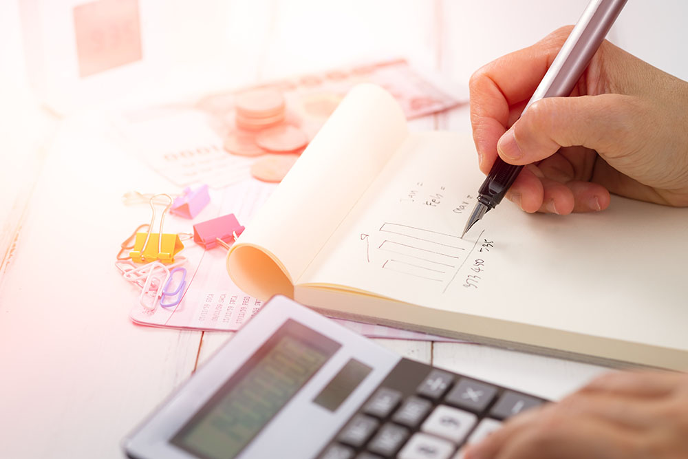 Uma pessoa anotando em um bloco de notas com uma calculadora ao lado. Imagem representa o cuidado com os custos do escritório