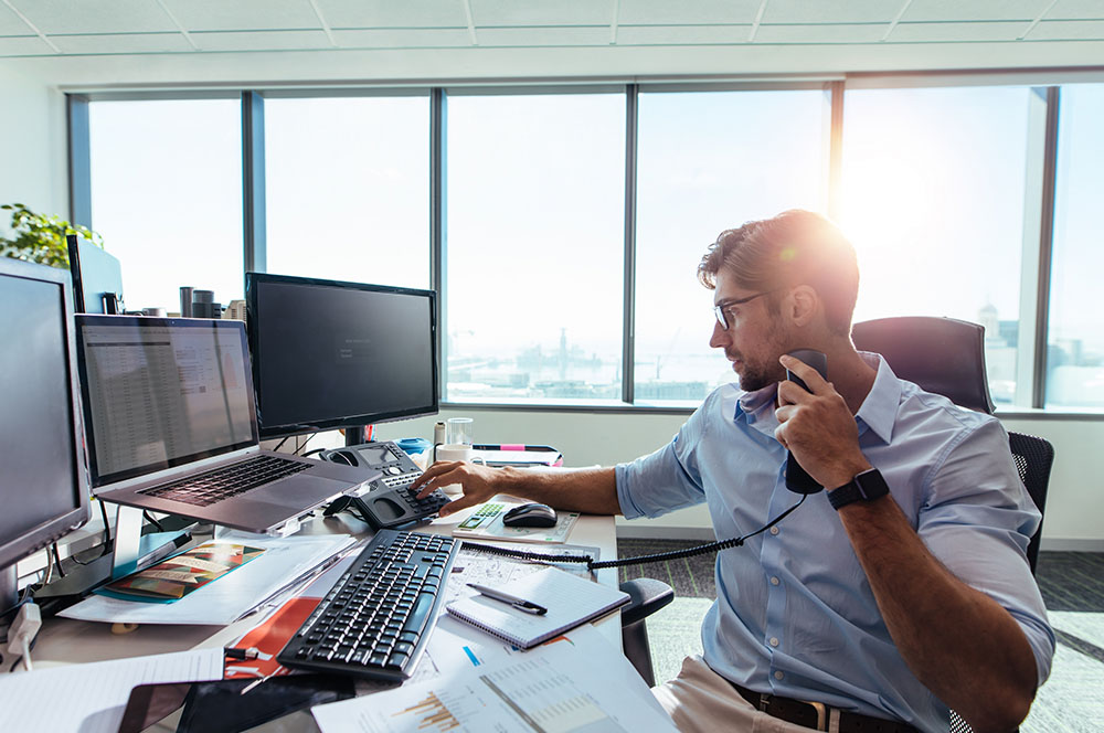 Um homem usando o telefone enquanto mexe em um computador em um escritório de advocacia com vista panorâmica. Foto está relacionada com Atendimento jurídico