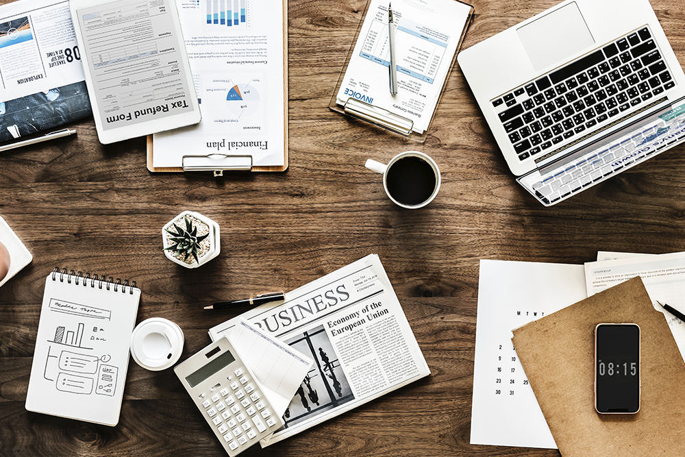 Foto de uma mesa com diversos elementos sobre ela. Gráficos, planilhas, jornal, notebook, bloco de notas, etc. Demonstra a necessidade de um Fluxo de trabalho na advocacia trabalho em escritórios corporativos