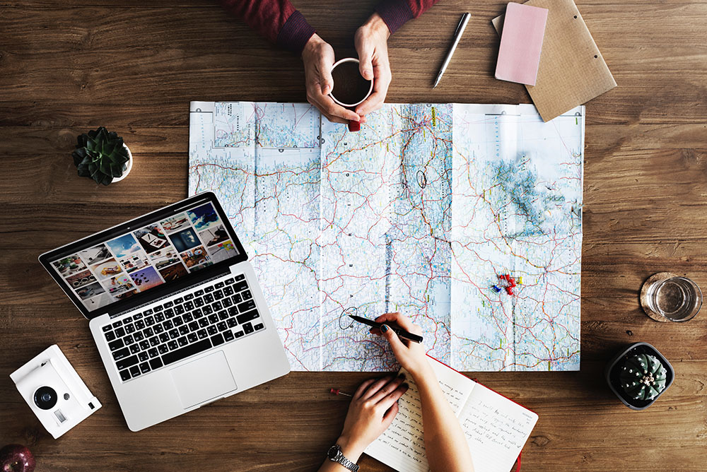 Uma foto aérea de uma mesa. Sobre ela, um notebook, blocos e caneta para anotação, e duas pessoas traçando rota em um mapa. representa a expansão geográfica permitida pelo Atendimento jurídico digital