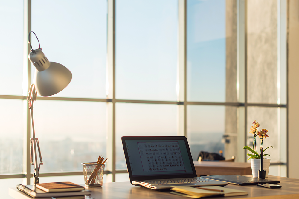 Um notebook e um abajur sobre um a mesa em frente a uma mesa com vista panorâmica. Representa a possibilidade de home office na advocacia