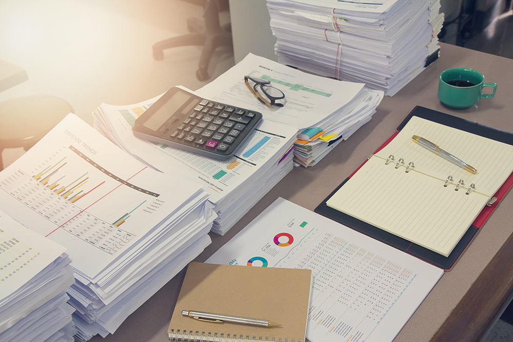 Uma mesa com diversas pilhas de relatórios e pastas, além de uma calculadora. Representa análises necessárias para identificar prejuízos no escritório