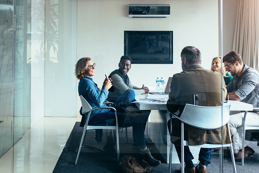 Três advogados e duas advogadas conversando e sorrindo sentados à uma mesa em um escritório bastante futuristas, com paredes de vidro. Simboliza o uso da tecnologia para qualificação da equipe.