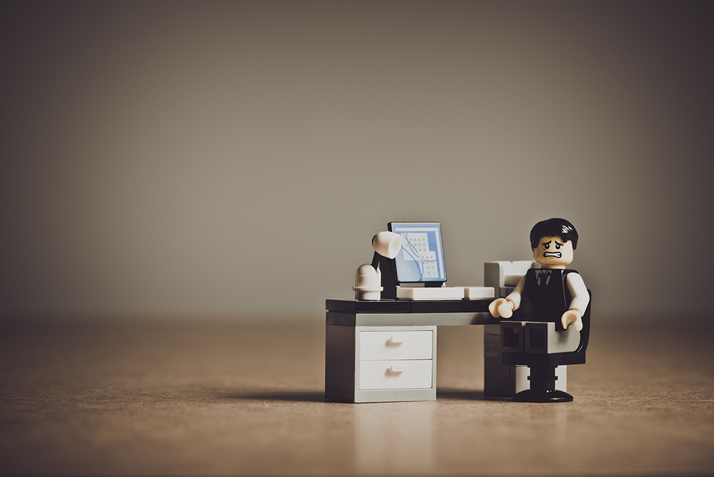 Foto de um bonequinho lego com semblante preocupado e usando roupas formais sentado em frente a uma escrivaninha com um computador.