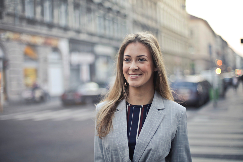Uma advogada vestida formalmente e com elegância, caminhando sorridente pelas ruas de uma cidade bonita. Representa o advogado bem-sucedido